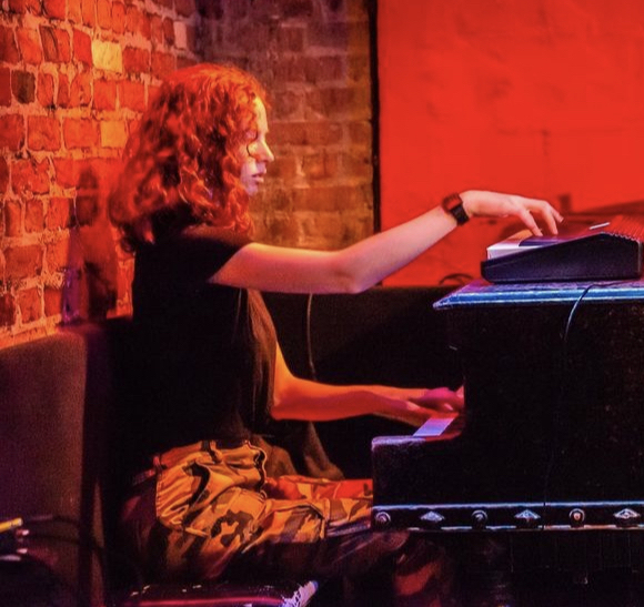 Julian is sitting at a grand piano with a keyboard on top of the lid. They are wearing a black shirt and orange pants.